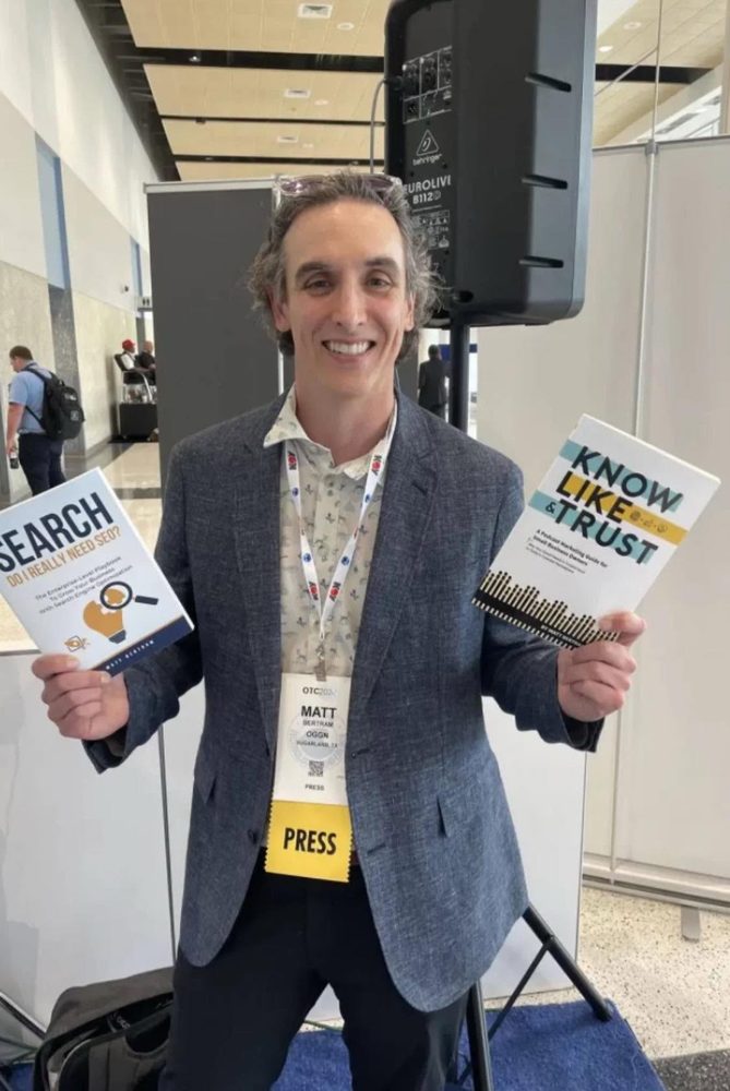 Matthew Bertram holding his books with a press badge at OTC 2026